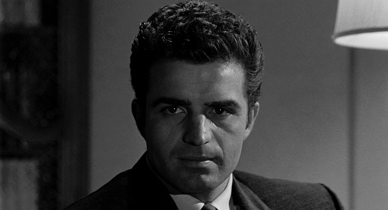 Murder By Contract (1958) movie still: closeup — An interior space features a man positioned against a dimly lit background. He faces the camera…