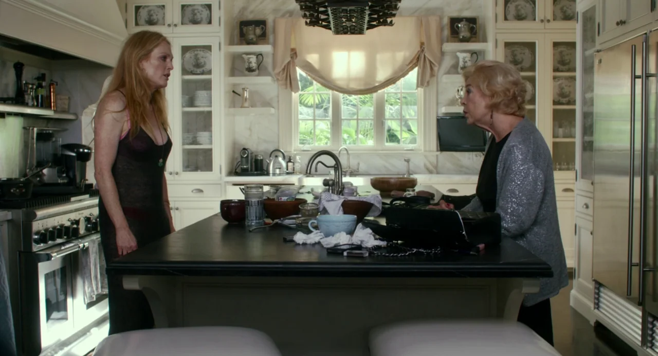 Maps to the Stars (2014) movie still: medium — A high-end modern kitchen with white cabinetry and a black stone island. Two women stand at the…
