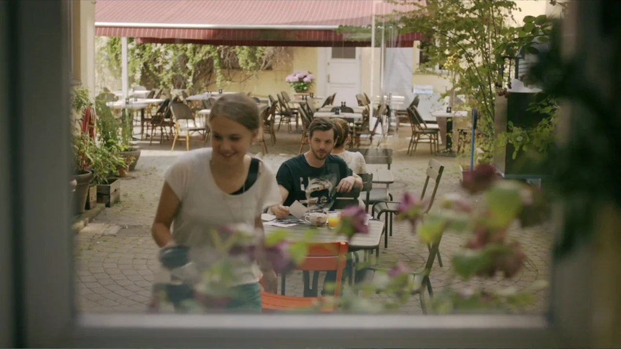 Copenhagen (2002) movie still: medium — An outdoor cafe terrace with multiple tables sits under a red fabric awning. A woman walks through the frame…