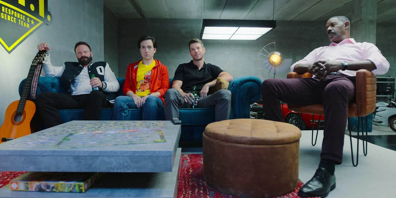 Sniper: G.R.I.T. (2023) movie still: wide — A modern garage with concrete walls serves as a communal seating area with furniture. Four men sit on a…