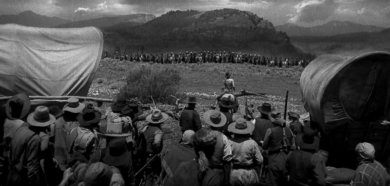 The Big Trail (1930) movie still: wide — A group of settlers with covered wagons faces an open plain in the West. A large line of riders on…