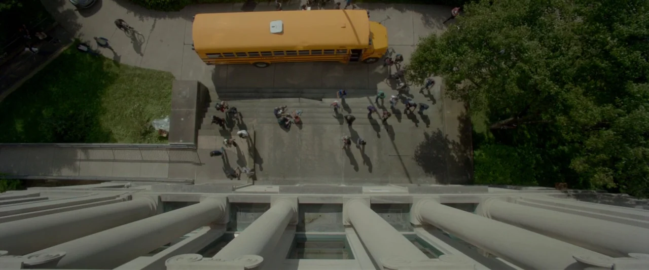 Me and Earl and the Dying Girl (2015) movie still: wide — A top-down view shows a yellow school bus parked before a stone building entrance. Groups…
