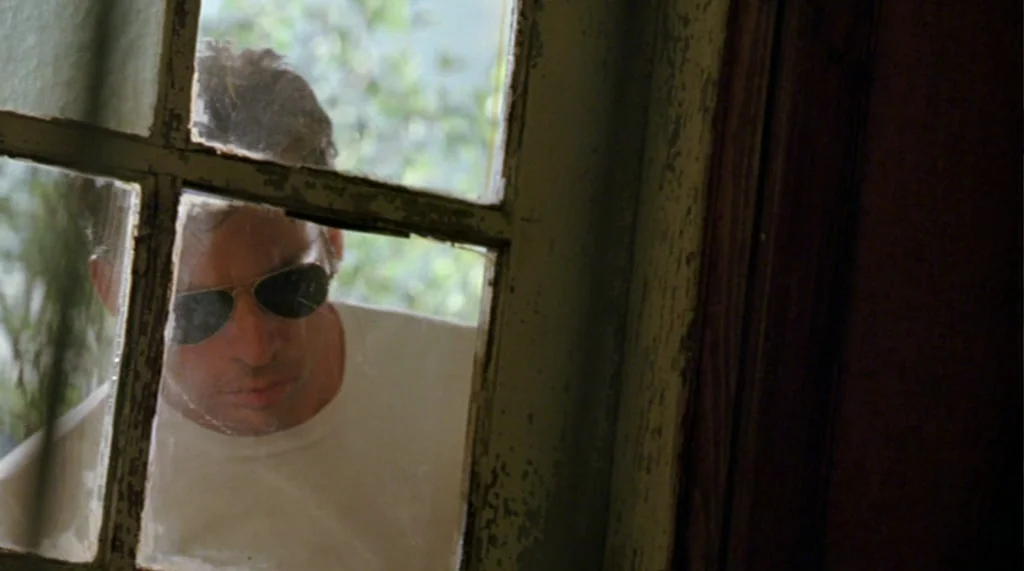 Bug movie still: closeup — A man peers through a weathered wooden frame window into a dimly lit room. The window pane is divided by multiple muntins…