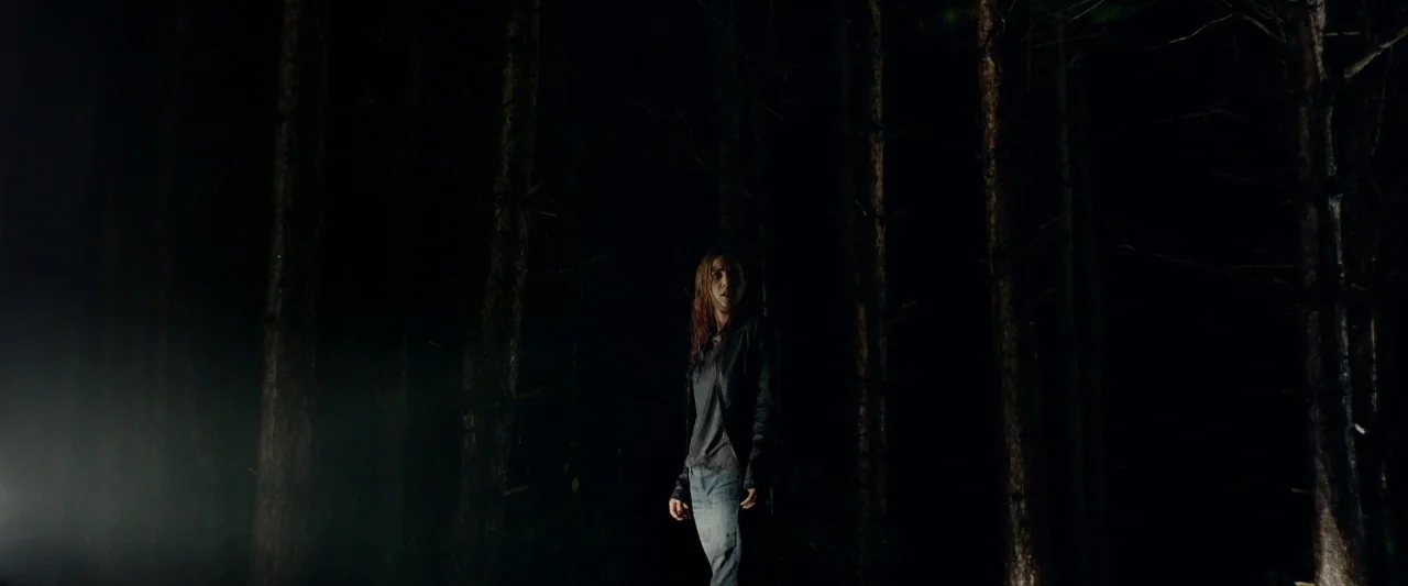 The Monster (2016) movie still: wide — A dense forest at night is shrouded in darkness with tall tree trunks visible. A young woman stands alone in…