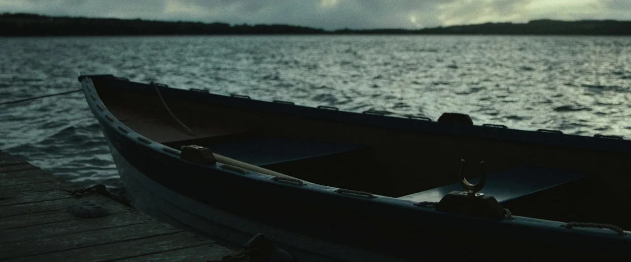 The Night House (2020) movie still: medium — A wooden rowboat is tied to a weathered plank dock on a grey lake. The water is textured with small…