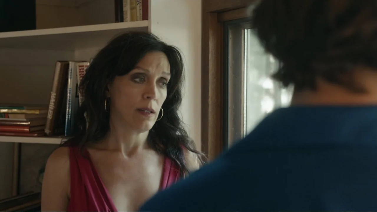 Toss It (2019) movie still: medium — A book-lined interior space features a woman standing by a wooden window frame. The woman speaks to a man seen…