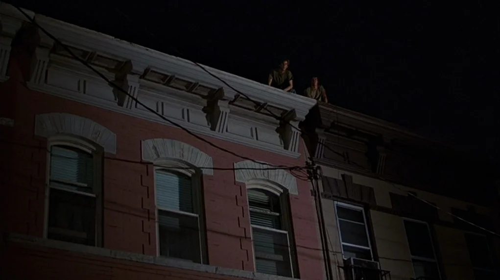 The Warriors (1979) movie still: wide — Two brick apartment buildings are lit at night from a low-angle street source. Two people are positioned on…