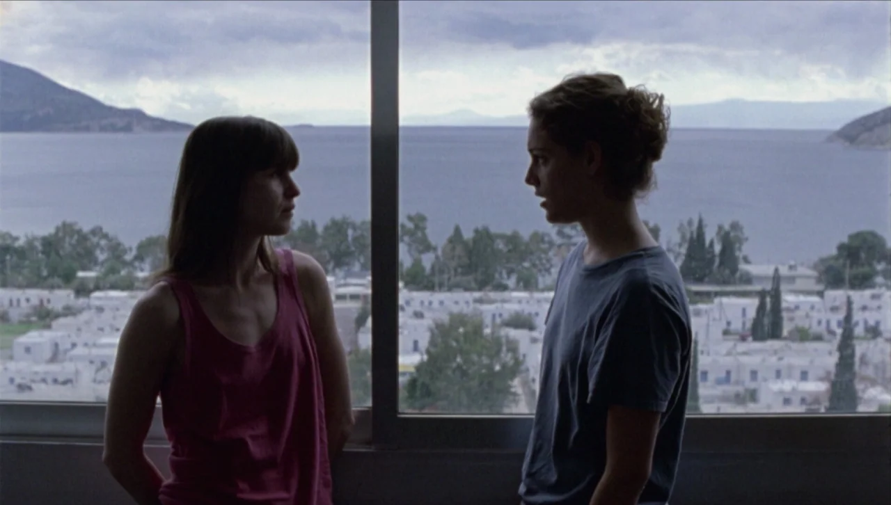 Attenberg (2010) movie still: medium — An interior hotel room features a large window overlooking a coastal village and water. Two women stand in…