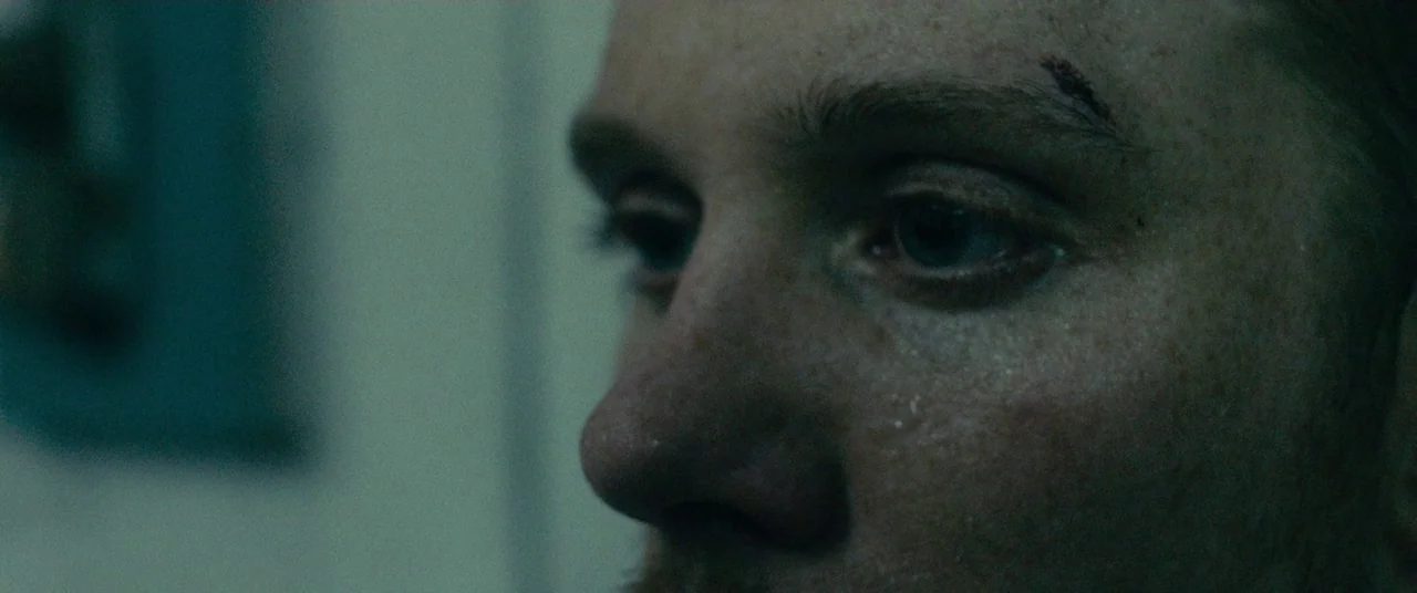 A Prayer Before Dawn (2017) movie still: closeup — A dark, dimly lit indoor space with muted blue and greenish tones. A man with a cut on his brow…