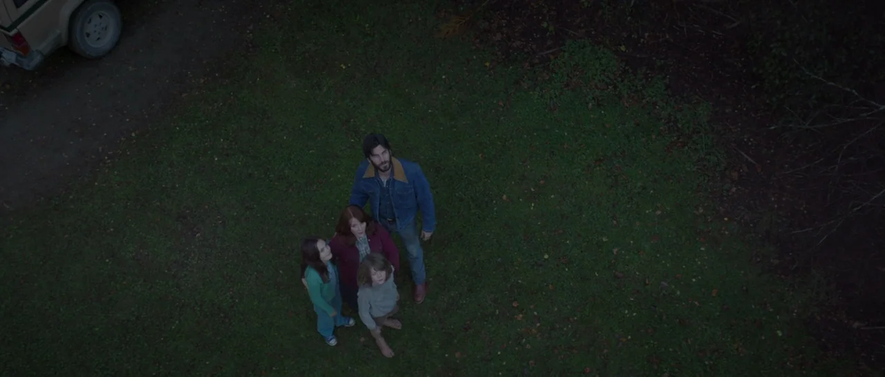 Pete’s Dragon (2016) movie still: wide — An overhead view of a family of four standing on a grassy area outdoors. They look upward toward the camera…