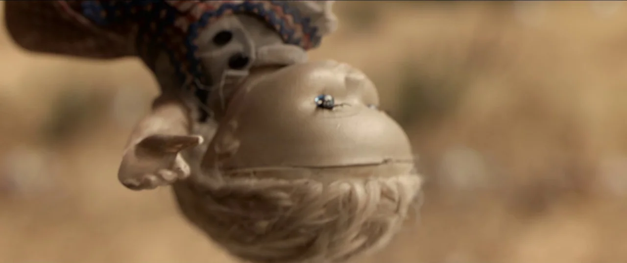 His House (2020) movie still: closeup — A plastic doll hangs suspended upside down against a blurred out-of-focus earth landscape. The doll features…