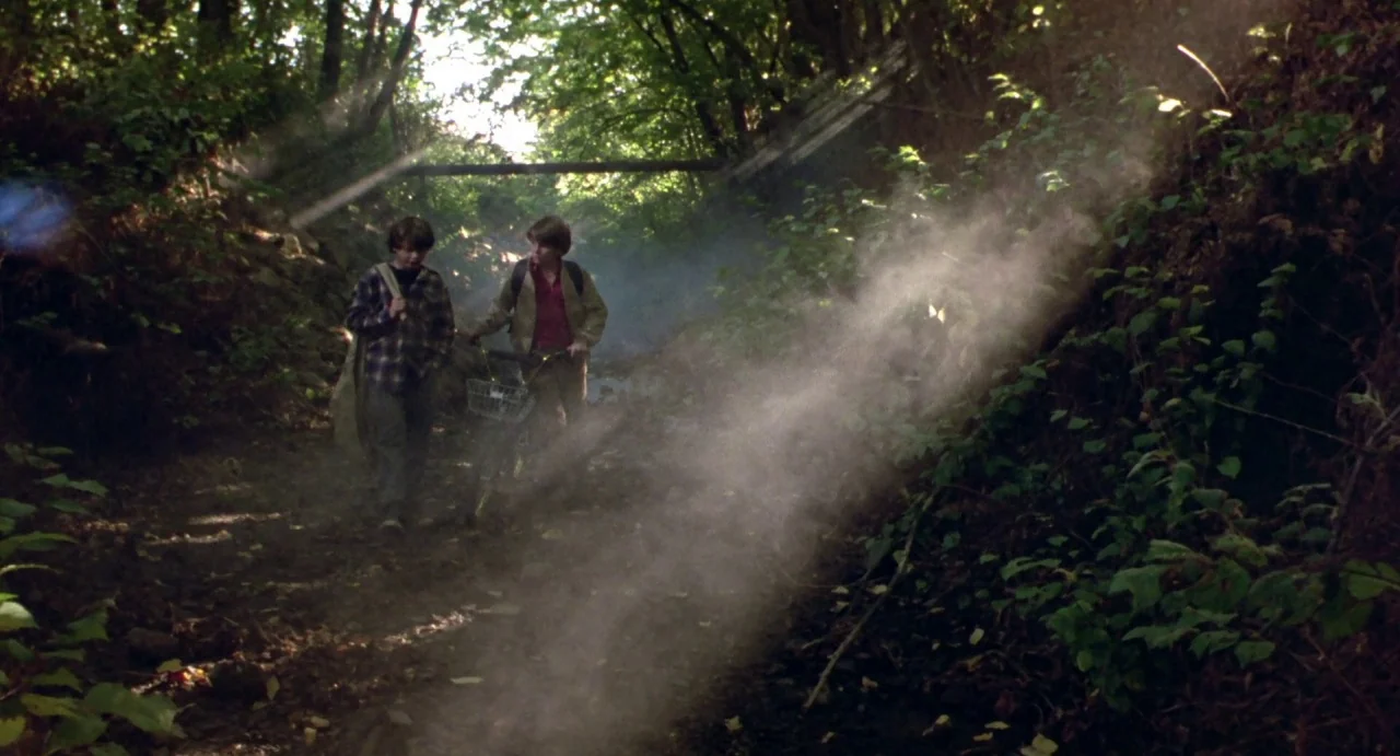 Explorers (1985) movie still: wide — A narrow dirt path through a dense wooded area with sunbeams cutting through haze. Two teenage boys walk slowly…