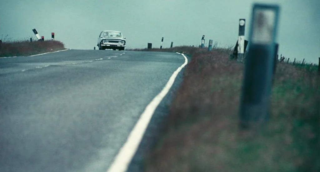The Damned United (2009) movie still: wide — A rural asphalt road curves gently upward under a grey, overcast sky. A single vintage sedan travels…