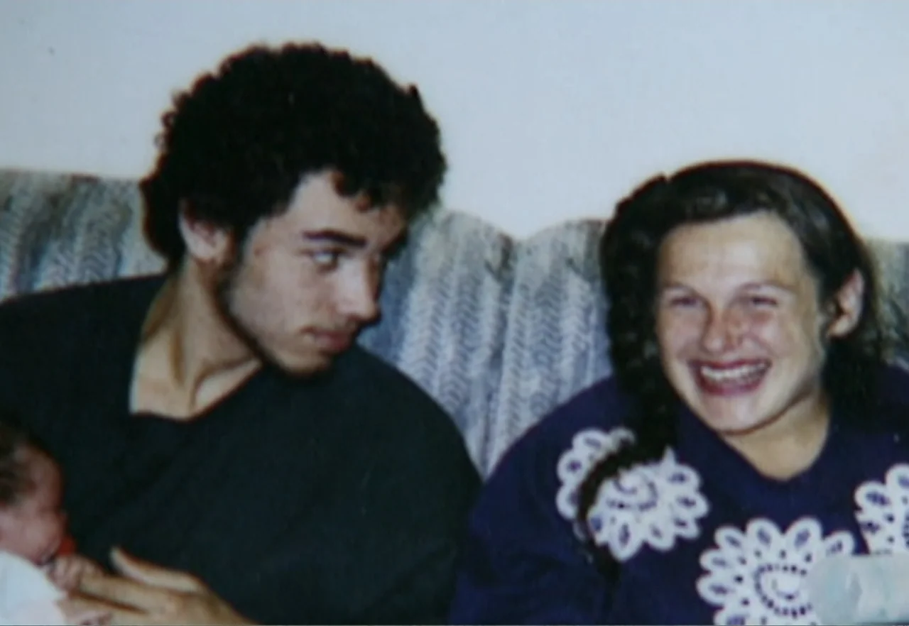 Broken Child (2000) movie still: medium — A domestic interior with a grey upholstered sofa against a plain white wall. A young man and woman sit…