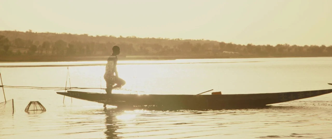 Timbuktu (2014) movie still: wide — A vast calm lake is bathed in golden light during the late afternoon. A lone man stands inside a slim wooden…
