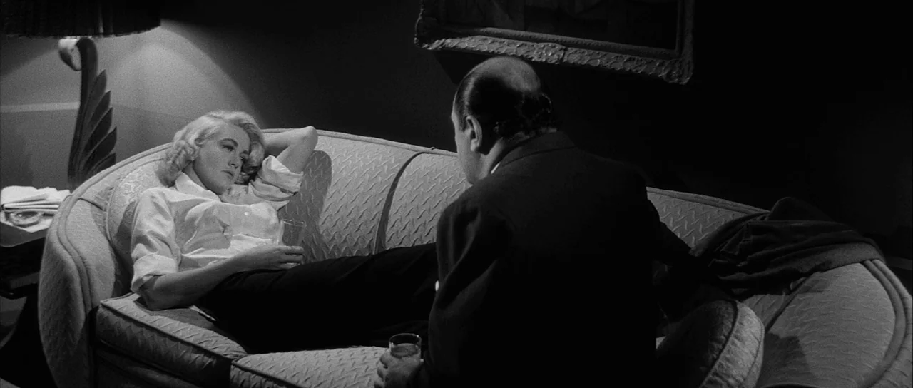 The Tarnished Angels (1957) movie still: medium — A dimly lit room features a curved sofa and a table lamp with an ornate base. A woman rests on the…