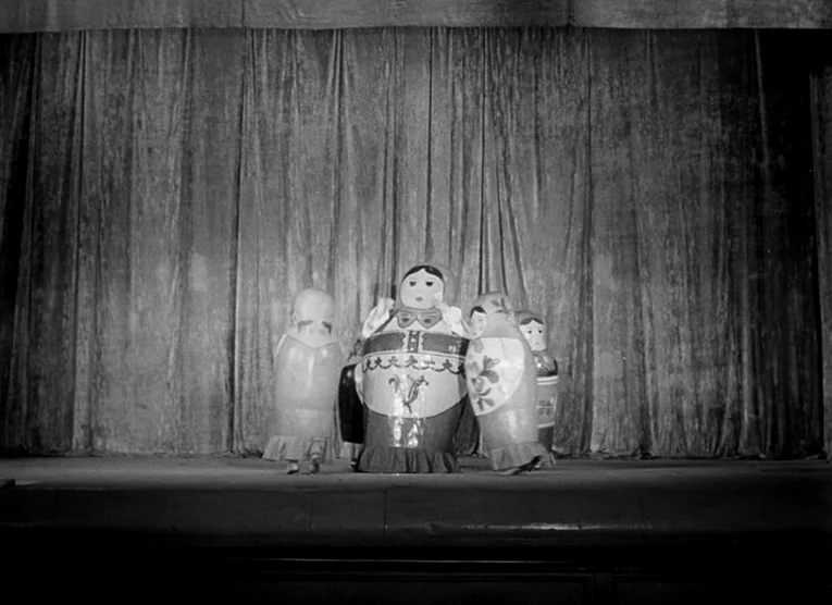 Wings (1966) movie still: wide — Four large, cylindrical dolls dressed in Russian folk-style costumes stand on a wooden stage. A heavy, pleated…