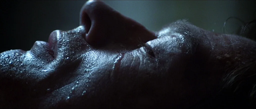 Alien (1979) movie still: closeup — A dark space focuses on the profile of a man's face. Moisture coats the skin as he remains still with closed eyes.