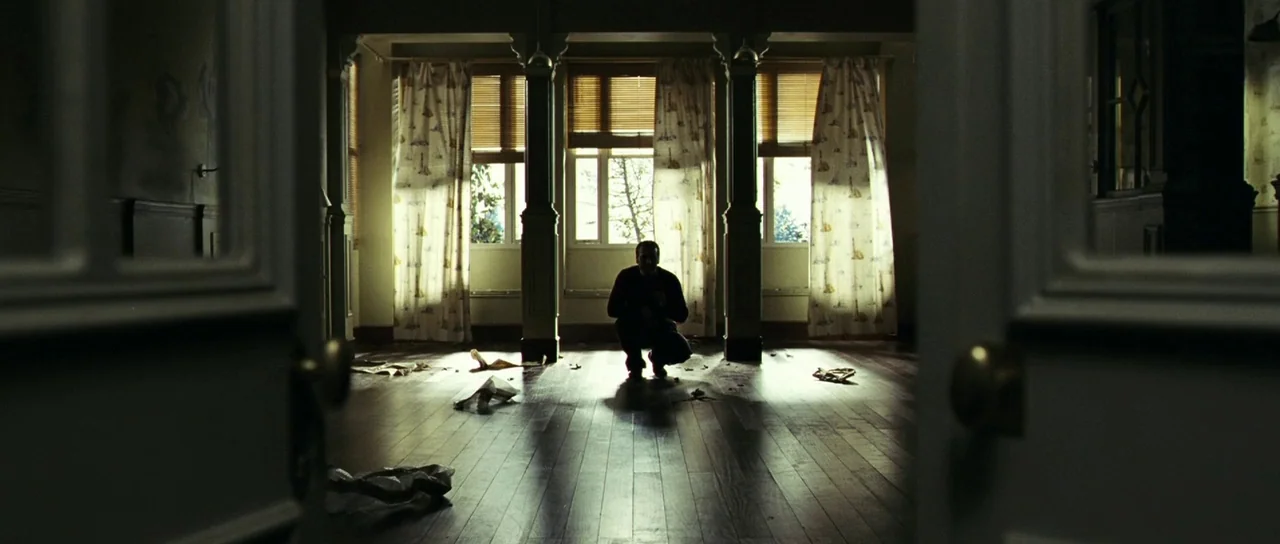 The Orphanage (2007) movie still: wide — A spacious room features polished wooden floors and large windows covered by patterned curtains. A man is…