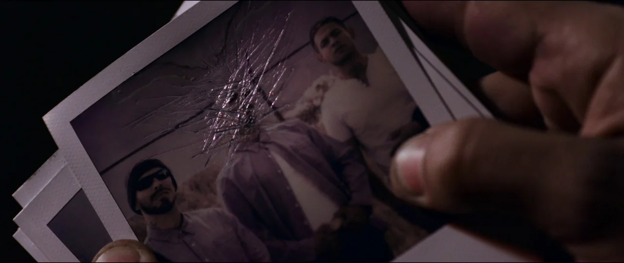 El Chicano (2018) movie still: closeup — A close-up view of a hand holding a photograph with a shattered surface texture. Three men are visible in…
