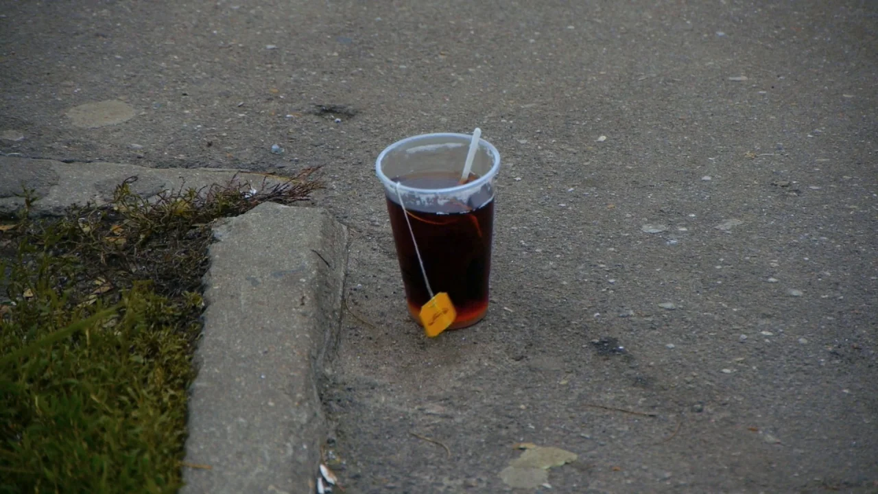 Counting (2015) movie still: closeup — A clear plastic cup containing dark tea sits on a textured grey asphalt surface. A yellow tea bag tag hangs…