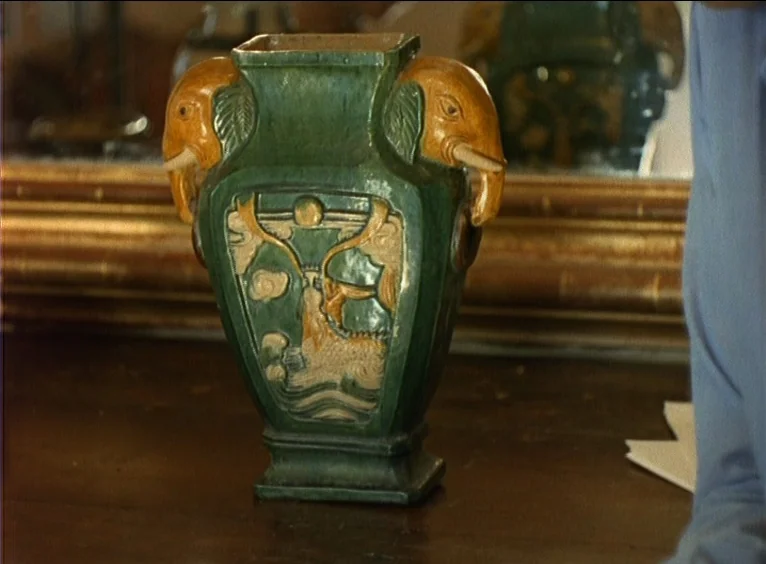 La Collectionneuse (1967) movie still: closeup — A dark wooden surface holds an ornate green ceramic vase with elephant-shaped handles. The vase…