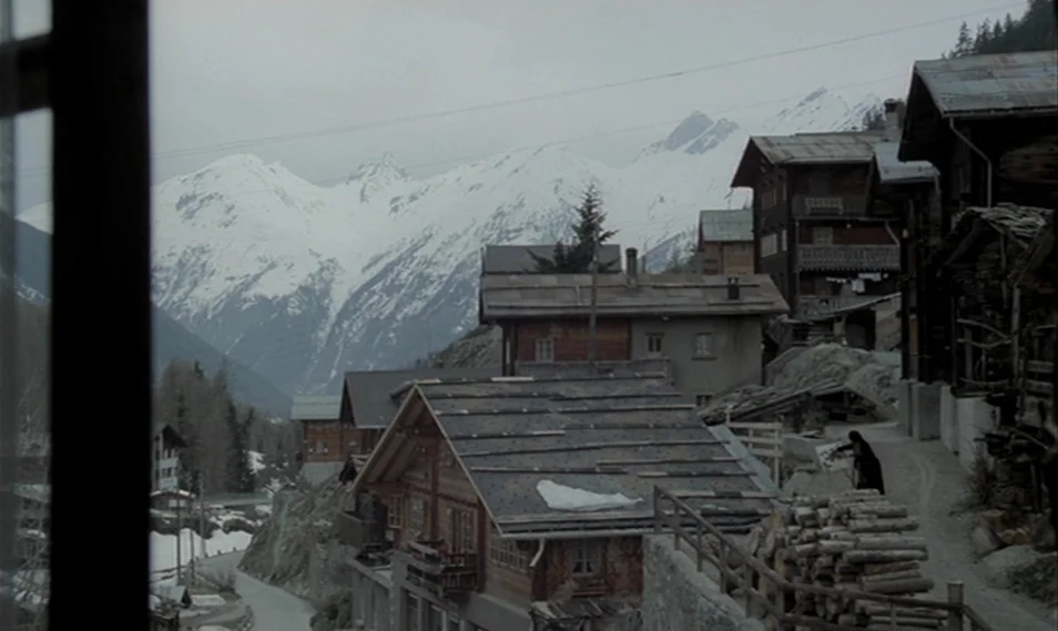 Despair (1978) movie still: establishing — A quiet village nestled in a valley with dark wooden houses and snow-capped peaks. An overcast sky looms…