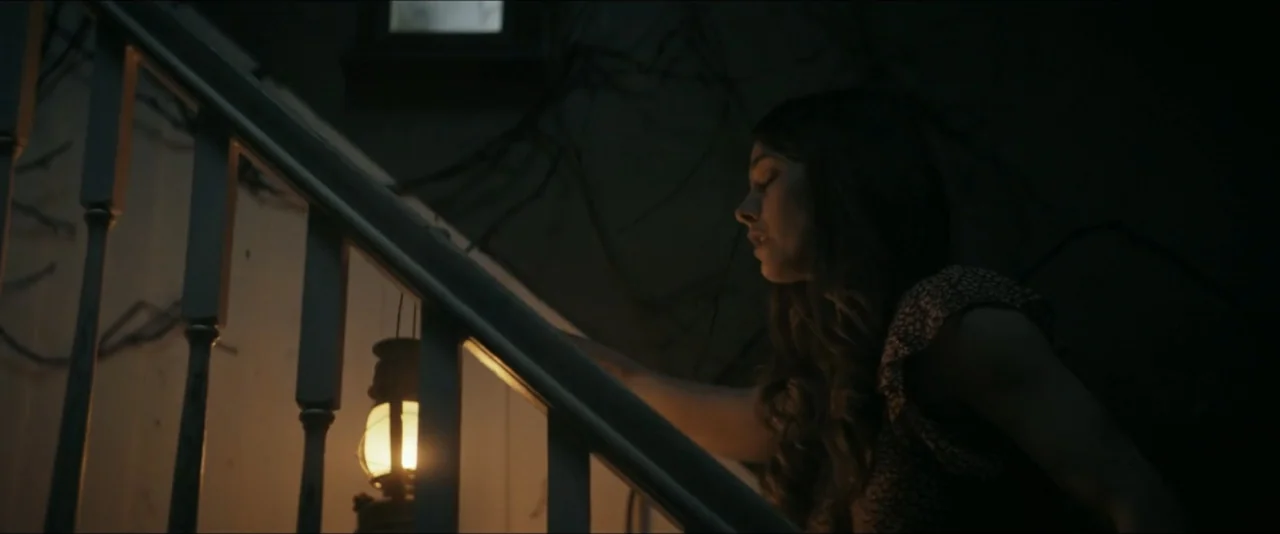 It Feeds (2025) movie still: medium — A wooden outdoor staircase with a dark wall covered in vines at night. A woman in a floral blouse ascends the…