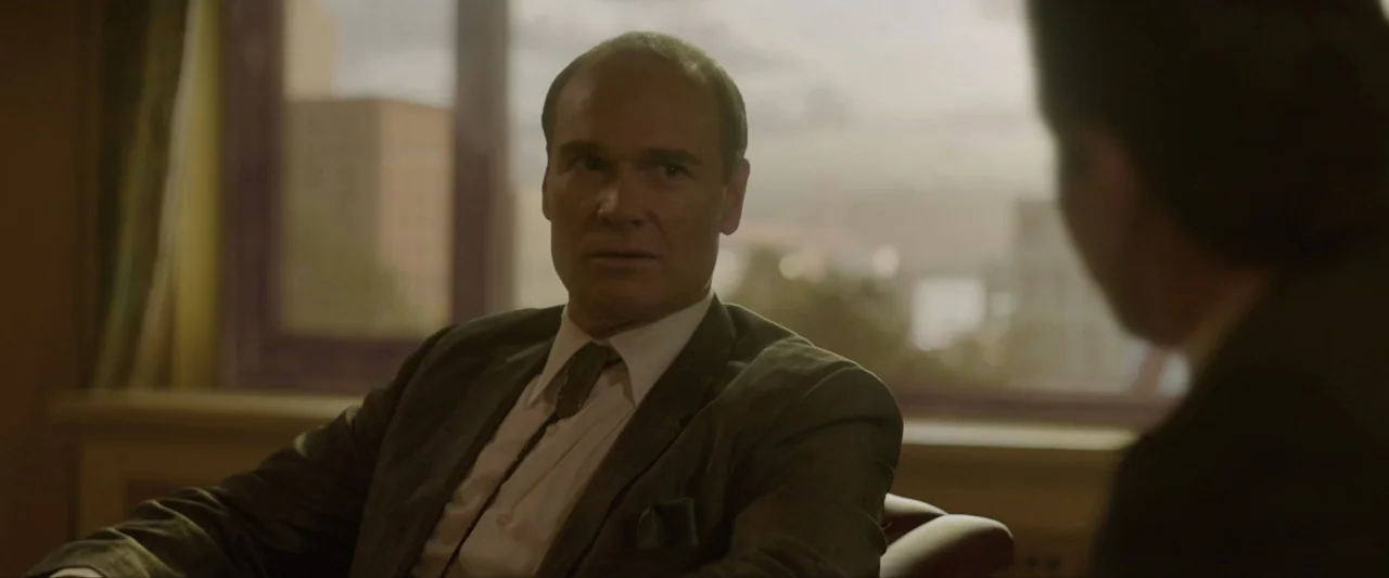 Reagan (2024) movie still: medium — An interior office space features a large window overlooking an indistinct urban landscape. A man in a grey suit…