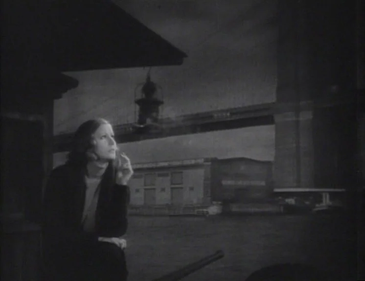 Anna Christie (1930) movie still: medium — A woman stands on a waterfront pier at night near a large bridge. In the background, industrial warehouse…