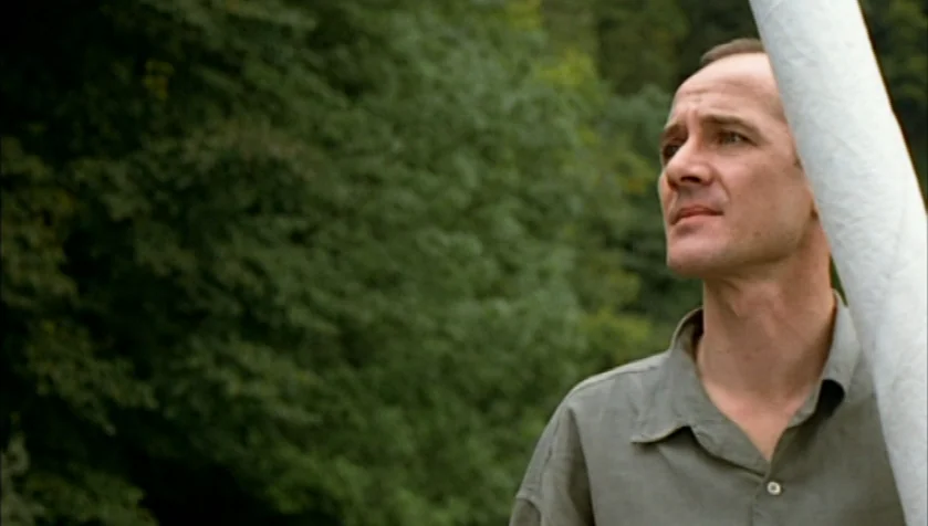 Funny Games (1997) movie still: medium — A man stands outdoors with lush green trees blurred in the background. He gazes off-camera to the left…