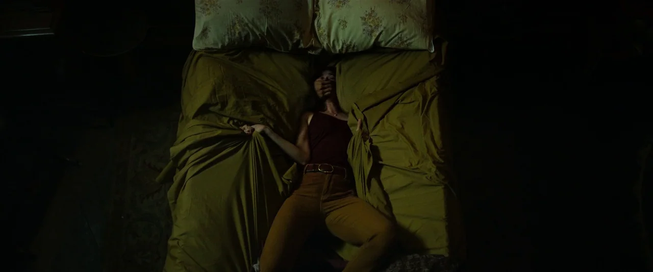 The Pope’s Exorcist (2023) movie still: medium — A dimly lit room features a person lying flat on an olive-colored bed. The subject clutches the…