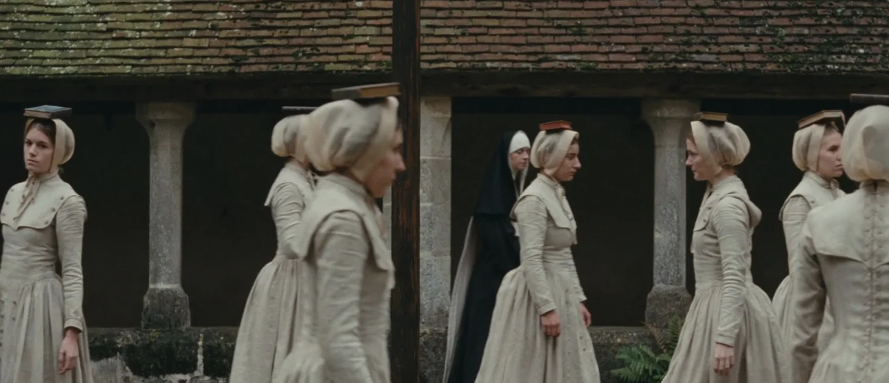 Madame Bovary movie still: wide — A stone cloister with tiled roofing provides the backdrop for a line of women. Several women in beige dresses walk…