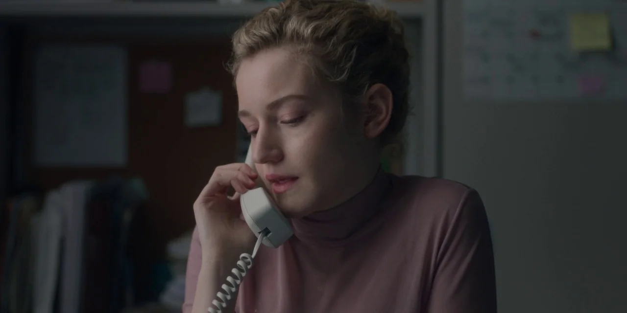 The Assistant (2019) movie still: closeup — An office environment with a desk area featuring blurred paperwork and post-it notes. A woman holds a…