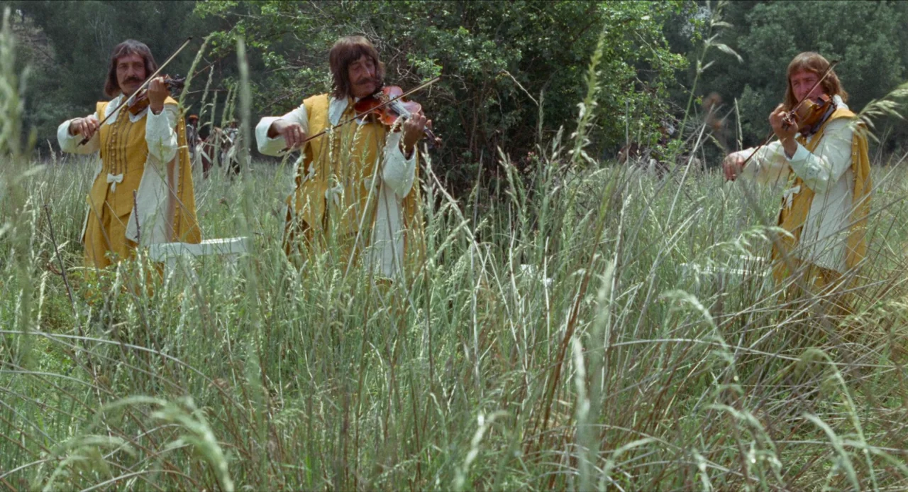 The Three Musketeers (1973) movie still: wide — A field of tall grass with three men playing violins in a row. They wear identical…