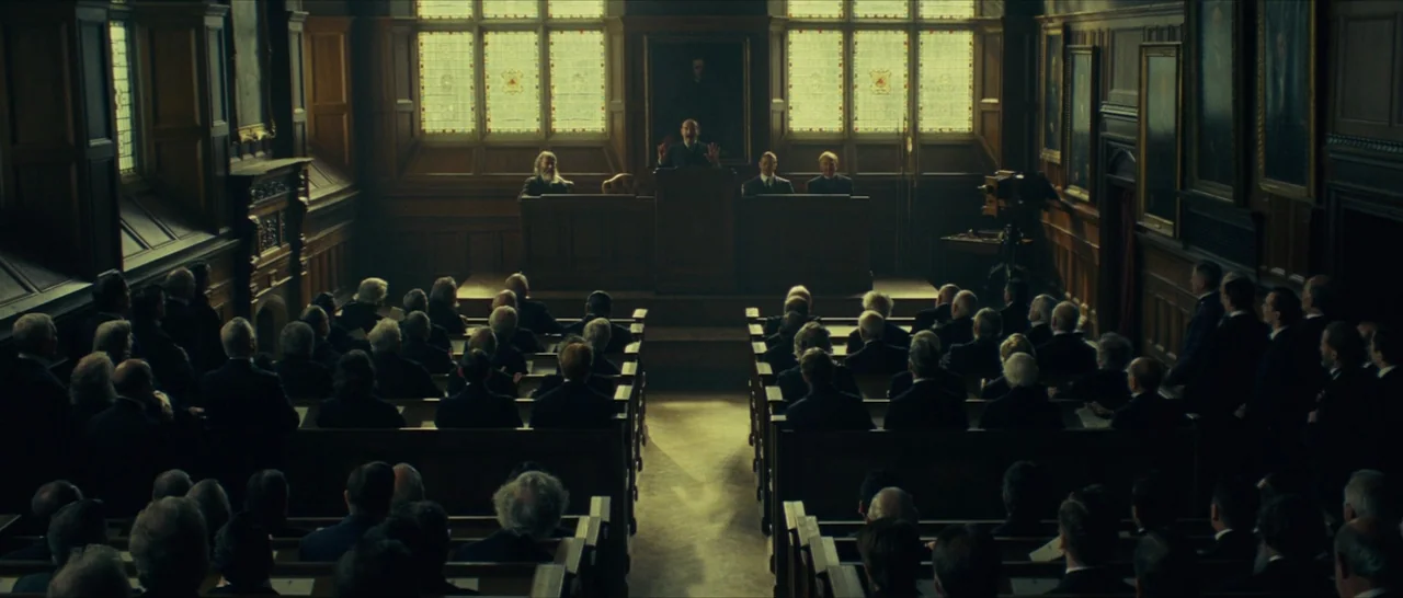 The Lost City of Z (2016) movie still: wide — A wood-paneled courtroom features rows of wooden pews facing a raised central podium. A man stands at…