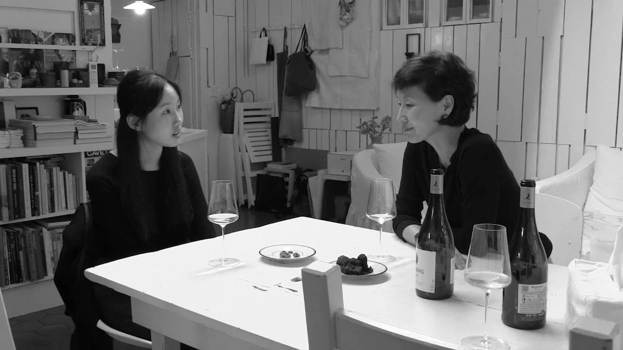 Walk Up (2022) movie still: medium — A minimalist white room with floor-to-ceiling bookshelves and wooden paneled walls. Two women sit at a white…