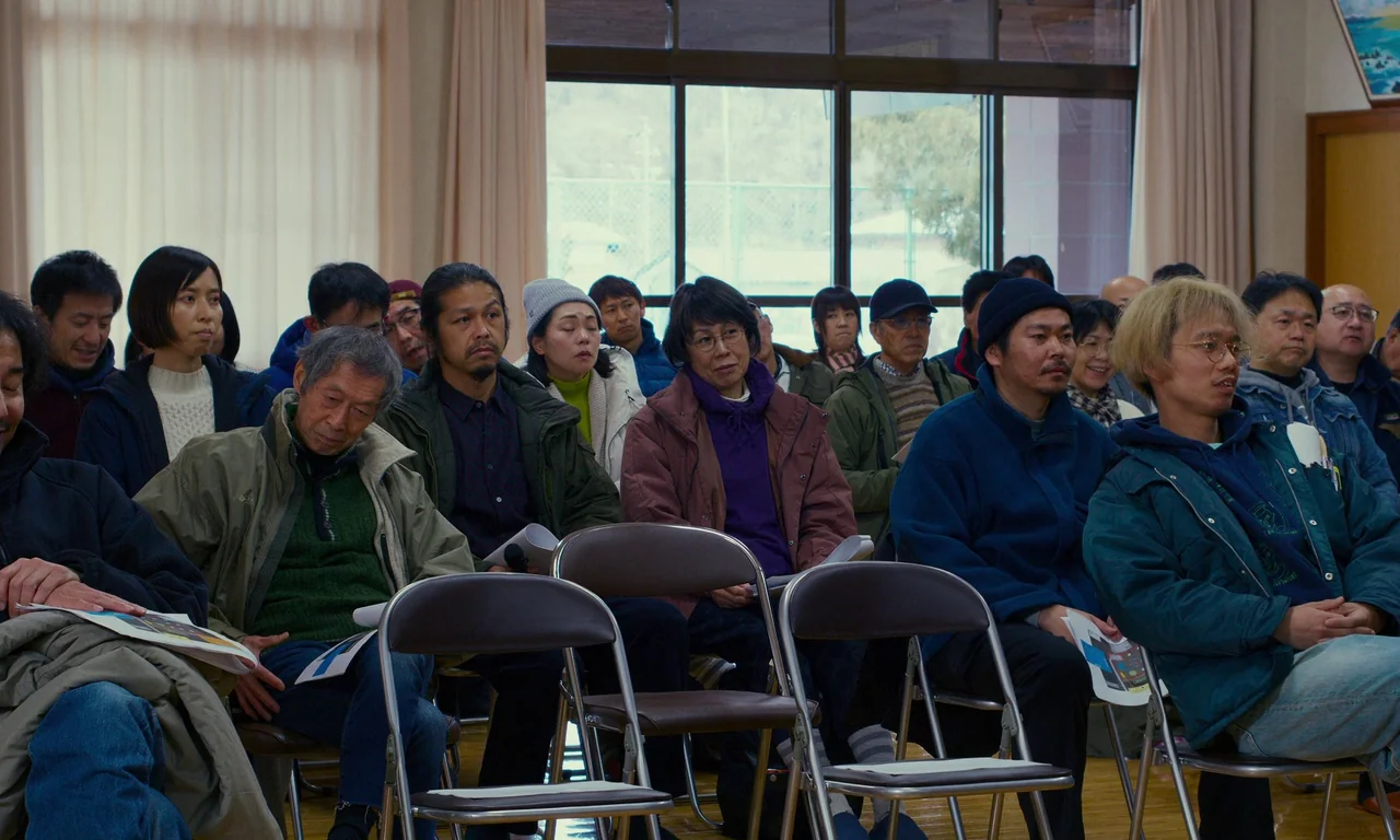 Evil Does Not Exist (2023) movie still: medium — A group of people sits in folding chairs inside a multipurpose community room. They hold documents…