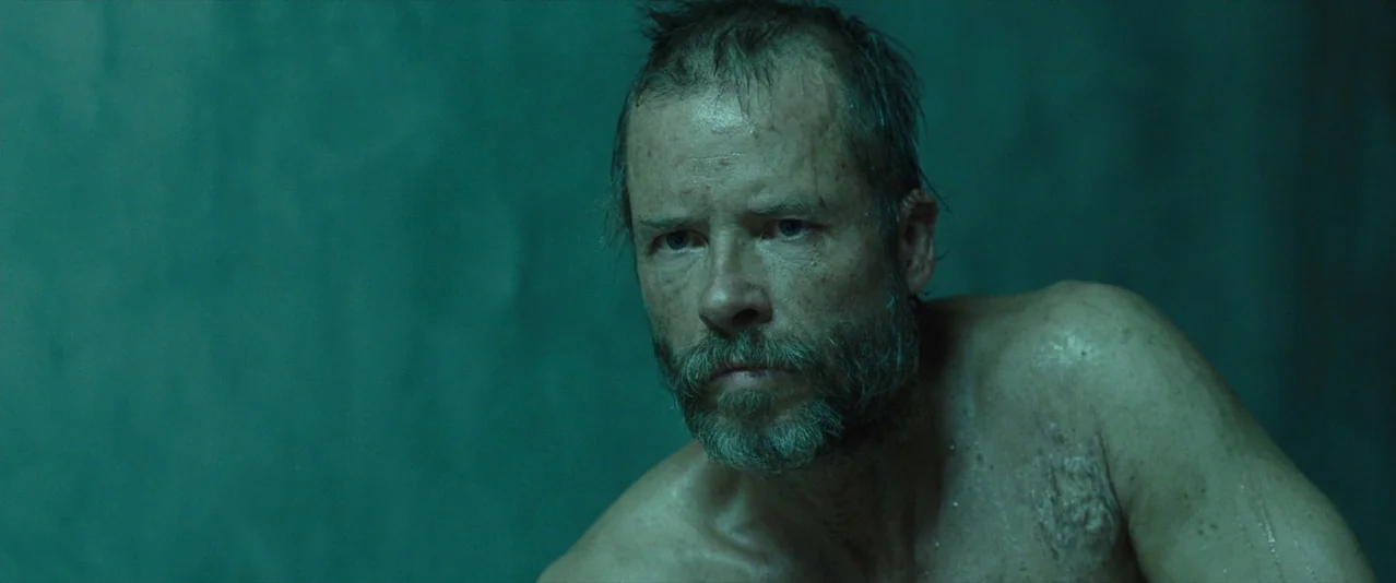 The Rover (2014) movie still: closeup — A man stands against a plain green wall in a dimly lit enclosure. His chest is damp and he faces slightly…