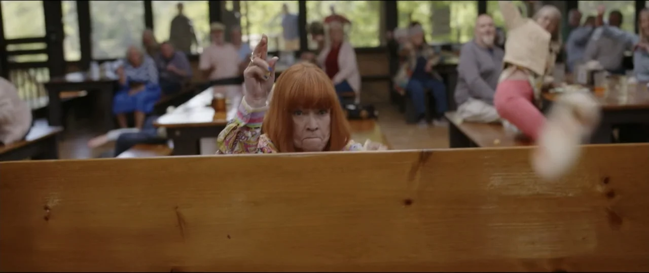 Summer Camp (2024) movie still: medium — A wooden mess hall features long benches with several seated people in the background. A woman in a…