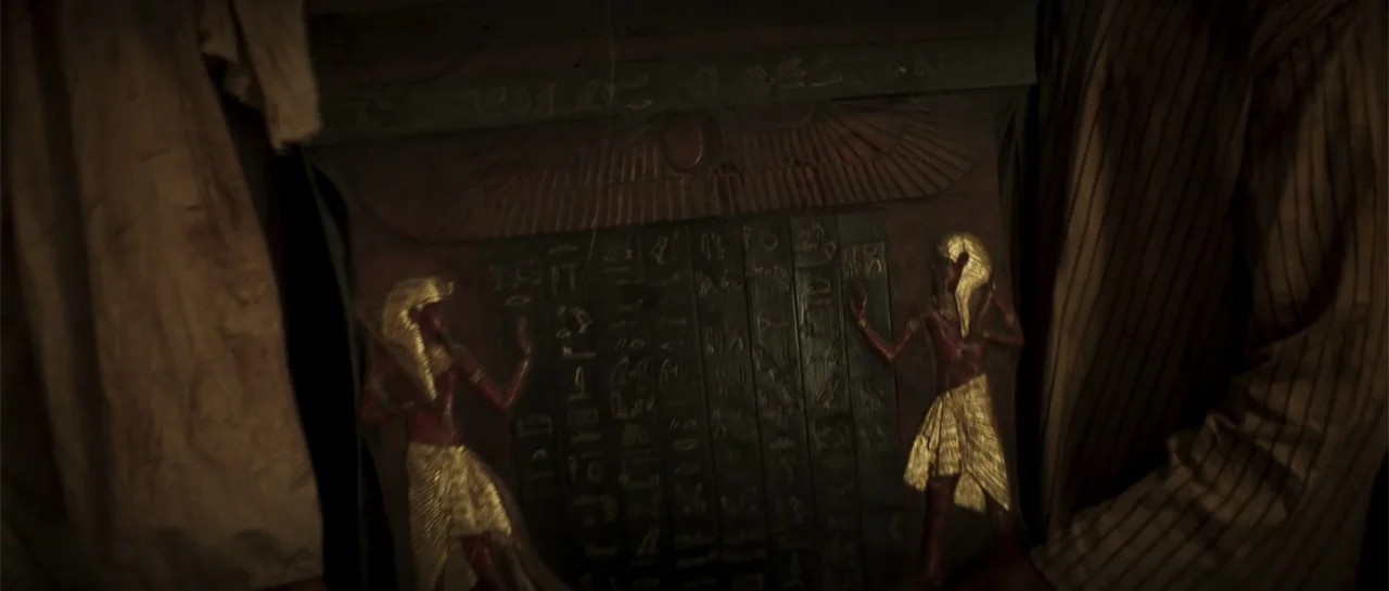 The Mummy (1999) movie still: closeup — A dimly lit room holds a dark wooden artifact featuring carved Egyptian figures and hieroglyphics. Two…