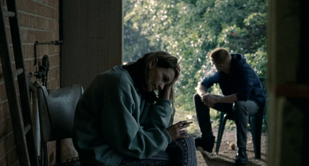 Snowtown (2011) movie still: medium — A woman sits in the foreground on an outdoor patio adjacent to a brick wall. A man sits on a plastic chair in…