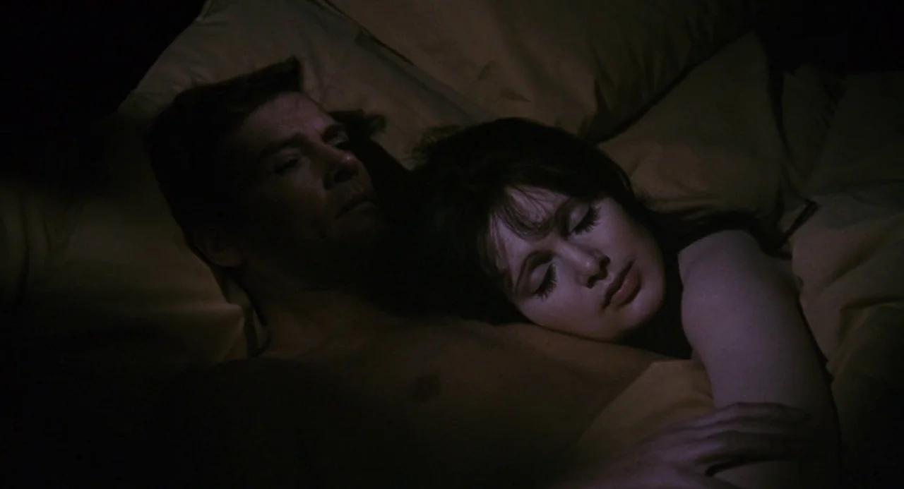 Live and Let Die (1973) movie still: medium — A dark bedroom shows two people resting together in a bed with yellow bedding. The couple lies with…