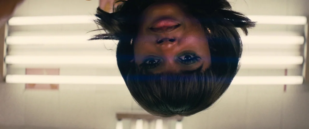 Licorice Pizza (2021) movie still: closeup — An interior space is illuminated by overhead fluorescent lighting bars arranged in rows. A woman looks…