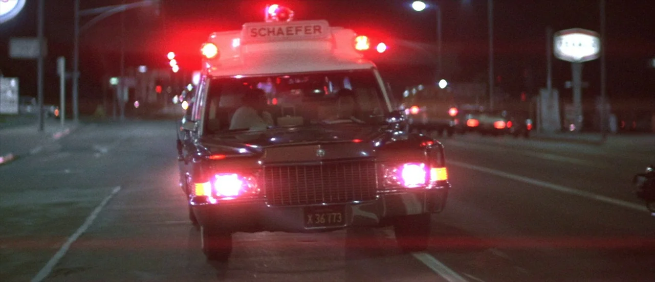 The Long Goodbye (1973) movie still: medium — An ambulance with flashing red lights travels down a dark asphalt road at night. The vehicle occupies…