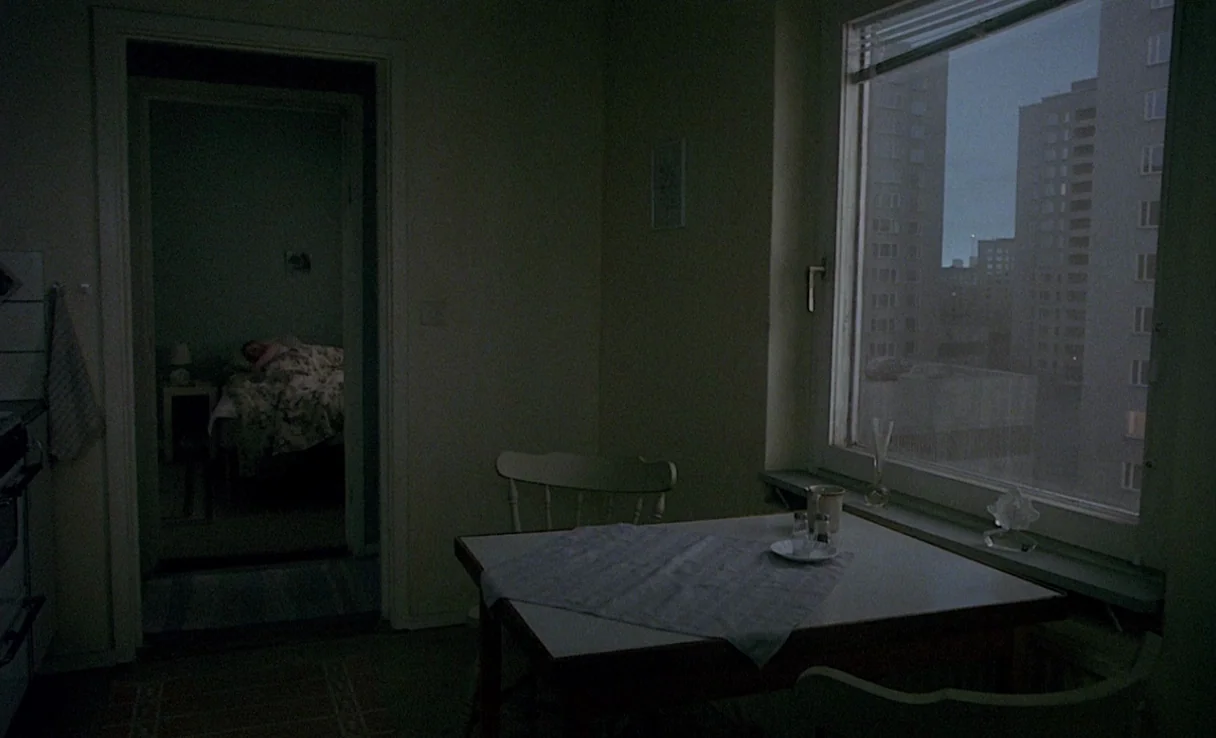 Songs from the Second Floor (2000) movie still: wide — A dimly lit apartment kitchen and dining area features a plain table with a cloth. A person…