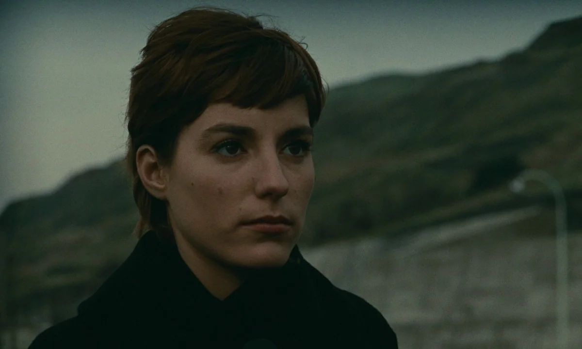 Muriel (1963) movie still: closeup — A woman stands in a desolate, hilly landscape under a dim, overcast sky. The subject looks off-camera while…