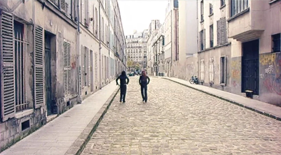 Scarlet Diva (2000) movie still: wide — A narrow residential cobblestone street is lined with weathered stone apartment buildings. Two individuals…