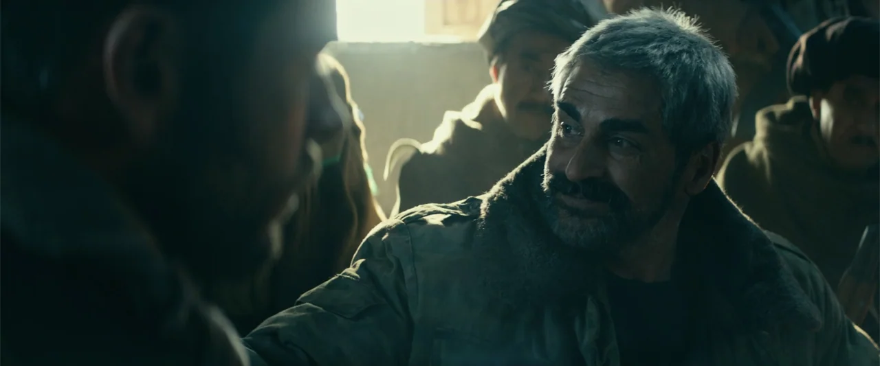 12 Strong (2018) movie still: medium — A dimly lit interior space with several men gathered together near a window. A middle-aged man in a military…