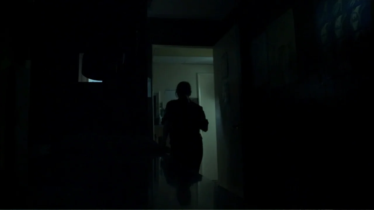 Abandoned Dead (2015) movie still: medium — A dark, unlit hallway leads to a brightly lit room visible through an open door. A person stands in the…
